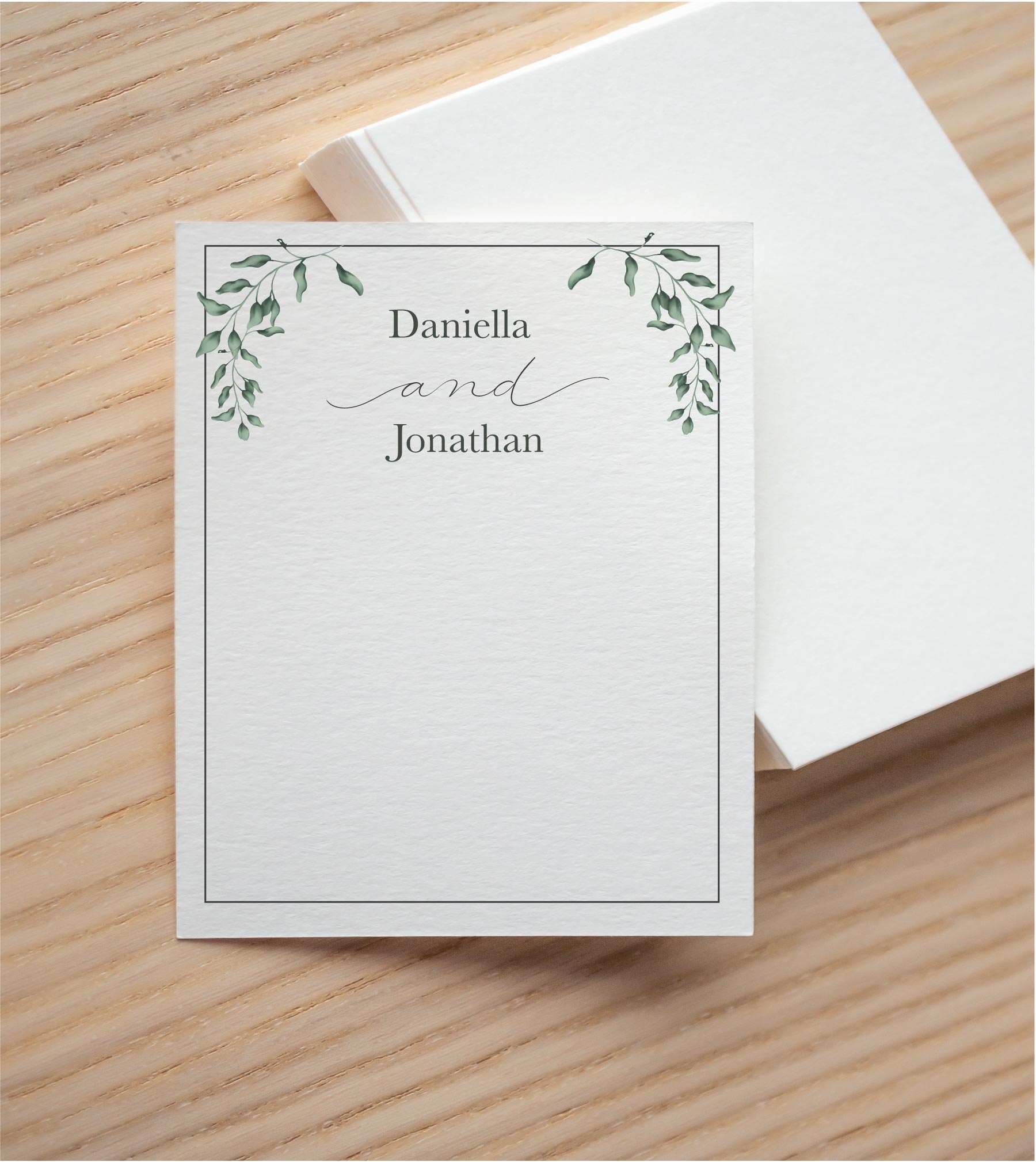 Personalized watercolor greenery thank you card with a couple's names in serif type and script "and," shown on a wood table with its box.